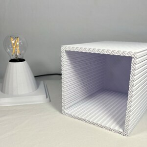 K&ouml;nnte beinhalten: Eine wei&szlig;e geometrische Lampe mit kegelf&ouml;rmigem Sockel und klarer Gl&uuml;hbirne. Daneben ein wei&szlig;er, gewellter W&uuml;rfel. Lampe und W&uuml;rfel stehen auf einer wei&szlig;en Oberfl&auml;che und erzeugen eine minimalistische &Auml;sthetik. Die Lampe ist eingesteckt.