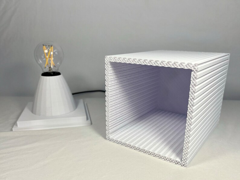 K&ouml;nnte beinhalten: Eine wei&szlig;e geometrische Lampe mit kegelf&ouml;rmigem Sockel und klarer Gl&uuml;hbirne. Daneben ein wei&szlig;er, gewellter W&uuml;rfel. Lampe und W&uuml;rfel stehen auf einer wei&szlig;en Oberfl&auml;che und erzeugen eine minimalistische &Auml;sthetik. Die Lampe ist eingesteckt.