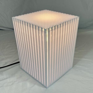 K&ouml;nnte beinhalten: Eine moderne, w&uuml;rfelf&ouml;rmige Lampe mit einer wei&szlig;en, gitterf&ouml;rmigen Oberseite und vertikalen, wei&szlig; gestreiften Seiten. Die Lampe ist von innen beleuchtet und hat ein schwarzes Netzkabel. Die Lampe ist etwa 30 cm hoch.