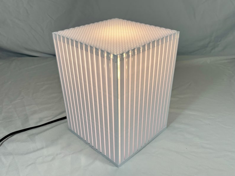 K&ouml;nnte beinhalten: Eine moderne, w&uuml;rfelf&ouml;rmige Lampe mit einer wei&szlig;en, gitterf&ouml;rmigen Oberseite und vertikalen, wei&szlig; gestreiften Seiten. Die Lampe ist von innen beleuchtet und hat ein schwarzes Netzkabel. Die Lampe ist etwa 30 cm hoch.