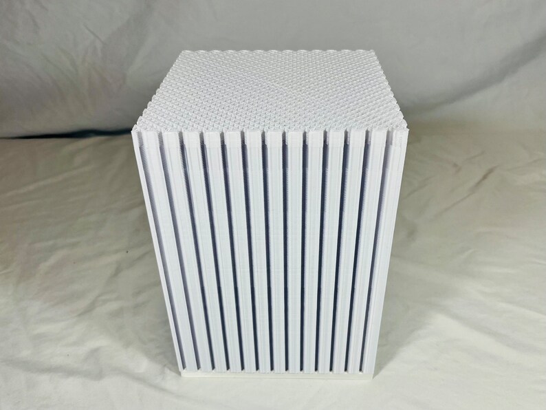 K&ouml;nnte beinhalten: Ein wei&szlig;es, w&uuml;rfelf&ouml;rmiges Objekt mit strukturierter Oberseite und vertikalen, geriffelten Seiten. Das Design weist ein sich wiederholendes Muster aus vertikalen Linien auf, das eine moderne, minimalistische &Auml;sthetik erzeugt. Das Objekt ist wahrscheinlich ein Dekorationsst&uuml;ck oder ein kleiner Tisch.