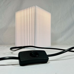 K&ouml;nnte beinhalten: Eine wei&szlig;e, rechteckige Lampe mit vertikalen Linien, die von innen beleuchtet wird. Ein schwarzes Kabel und ein Ein-/Ausschalter befinden sich im Vordergrund. Die Lampe ist ein modernes Design, geeignet f&uuml;r ein Schlaf- oder Wohnzimmer.