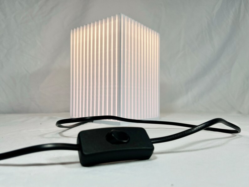K&ouml;nnte beinhalten: Eine wei&szlig;e, rechteckige Lampe mit vertikalen Linien, die von innen beleuchtet wird. Ein schwarzes Kabel und ein Ein-/Ausschalter befinden sich im Vordergrund. Die Lampe ist ein modernes Design, geeignet f&uuml;r ein Schlaf- oder Wohnzimmer.