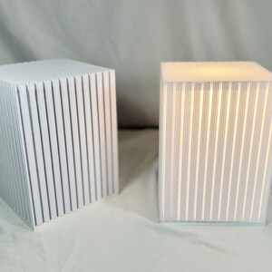 K&ouml;nnte beinhalten: Zwei wei&szlig;e, w&uuml;rfelf&ouml;rmige Lampen mit vertikalen Linien. Eine Lampe ist beleuchtet und strahlt ein warmes Leuchten durch die Linien aus. Die unbeleuchtete Lampe befindet sich links. Die Lampen stehen auf einer wei&szlig;en Oberfl&auml;che.