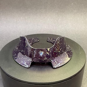 May include: A dark purple, speckled decorative collar with a glossy finish. The collar has a unique shape with cutouts and a small, iridescent gem. It sits on a black, circular base, against a dark background.
