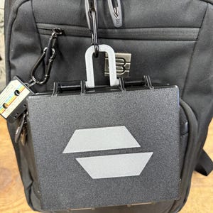 May include: A black briefcase-style case with a silver geometric design is attached to a black backpack. The case has a silver handle and is secured with a silver carabiner. A cassette tape keychain hangs from the backpack.