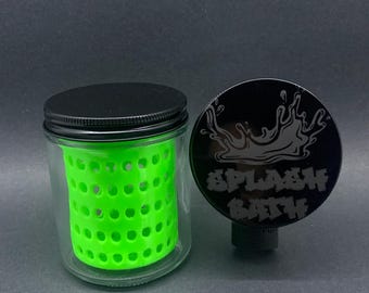 Peut inclure: Un bocal en verre transparent avec un couvercle noir et un insert perforé vert vif. À côté, un disque noir avec les mots "SPLASH BATH" et un motif d'éclaboussure.