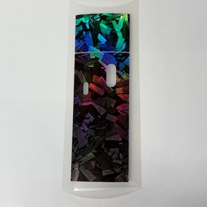 May include: A rectangular adhesive skin for an electronic device featuring a holographic, iridescent finish with a dark, fragmented, wood-like pattern. The sticker includes two precise cutouts for buttons or sensors and a clear protective backing.