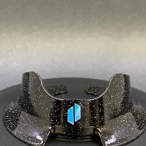 May include: A black, glitter-covered visor with a curved design and a blue logo. The visor has a wide brim and is designed to sit on the head. The logo features two vertical blue rectangles. The visor is on a black surface.