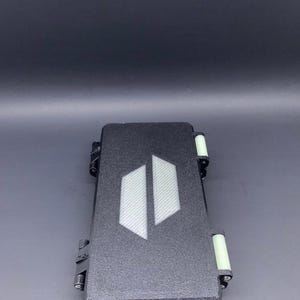 May include: A black rectangular case with a textured surface and a white geometric design on the lid. The case has hinges and clasps, and the ends have light green accents. The case is closed.