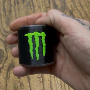 May include: A black cylindrical object with a neon green 'M' logo, held in a hand. The logo is the prominent feature, set against the black background. The object appears to be a small container or cup, possibly a shot glass.