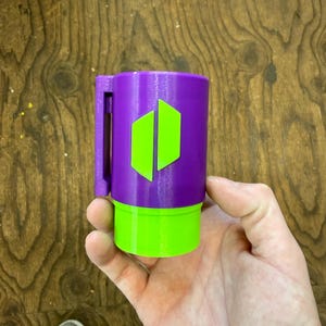 May include: A cylindrical mug with a handle, featuring a vibrant color scheme. The upper portion is a deep purple, while the base and a design element are bright green. The design on the mug appears to be two vertical green shapes.