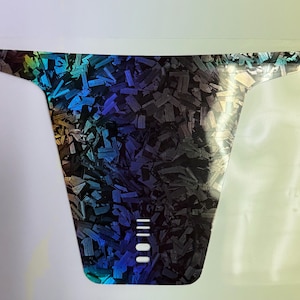 May include: A holographic sticker with a unique pattern of blue, green, and black shapes. The sticker has a distinctive shape with a wide top and a narrower bottom, and three white vertical lines.