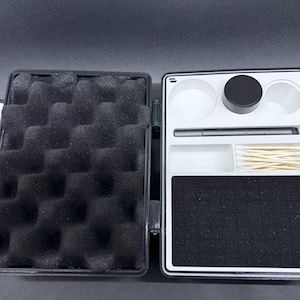 May include: An open black carrying case with a white interior. The left side has black foam padding. The right side has a white tray with a black container, two white containers, a pen, a small white tray, a small tray with cotton swabs, and a black felt pad.