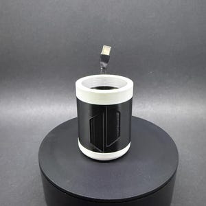 May include: A black and white cylindrical object with a USB cable extending from the top. The object has a white rim and a black body with a geometric design. It is sitting on a black circular base.
