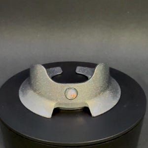 May include: A silver-colored, decorative object with a circular center and curved edges. The object has a glittery texture and a small, round, iridescent stone in the center. It sits on a black, circular base.