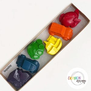 May include: A set of six crayons shaped like vehicles, including a plane, truck, boat, car, and two other vehicles. The crayons are arranged in a rainbow order in a white box with the text "Crayon Away" on the bottom left corner.