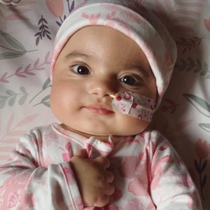 May include: A baby in a pink and white floral patterned onesie and hat. A medical tube is taped to the baby's face. The background features a floral design in pink and white.