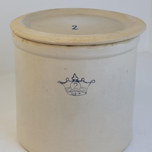 May include: A cylindrical off-white stoneware crock with a matching lid. The lid features a blue number 2. The front of the crock displays a blue stamped graphic of a crown with a number 2 inside and the text U.S.A. printed below the crown.