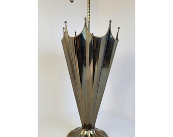 May include: A metallic umbrella stand with a brass-toned finish, shaped like an inverted umbrella with a curved handle at the top. The stand features a wide, circular base and a fluted, conical body with pointed edges along the rim.