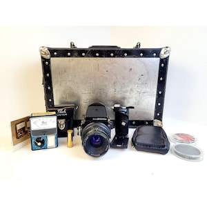 Puede incluir: Una cámara de formato medio Zenza Bronica negra con objetivo, un flashímetro en estuche negro, un pequeño cilindro dorado, una empuñadura negra, una funda de cuero negra, dos tapas de objetivo circulares y un maletín de transporte metálico.