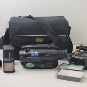 May include: A black Sony Handycam Video 8 camcorder with 12x zoom, accompanied by a black Aosta carrying bag, a battery pack, a lens attachment, a power adapter, a remote control, and two video cassettes, all arranged for a complete set.