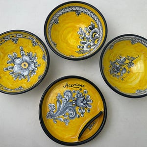 Può includere: Tre ciotole in ceramica gialla e un piatto abbinato con bordo nero. Ogni pezzo presenta motivi floreali grigi dipinti a mano. Il piatto include un piccolo scomparto laterale e la parola Aceitunas scritta in nero sulla superficie gialla.