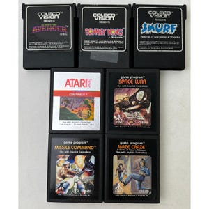 May include: A collection of seven vintage video game cartridges including three black ColecoVision titles: Cosmic Avenger, Donkey Kong, and Smurf. The four Atari 2600 cartridges feature Centipede, Space War, Missile Command, and Maze Craze labels.