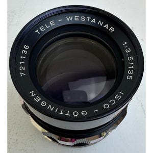 Puede incluir: Un objetivo de cámara negro Tele-Westanar con una especificación de apertura y distancia focal de 1:3,5/135. El elemento frontal es oscuro y el anillo exterior presenta el texto Tele-Westanar, Isco-Göttingen, 721136 y 1:3,5/135 en blanco.