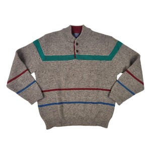 Puede incluir: Un suéter de manga larga en color beige jaspeado con cuello alto y dos botones en el cuello. El suéter presenta una franja horizontal de color verde azulado en el pecho y la parte superior de las mangas, con rayas rojas y azules más finas.