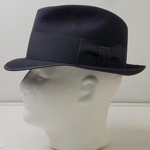 Könnte beinhalten: Ein dunkelblauer Fedora-Hut mit einem passenden dunkelblauen Ripsband und einer kleinen dekorativen Schleife an der Seite. Der Hut hat eine strukturierte Krone und eine leicht geschwungene Krempe mit einer glatten, gleichmäßigen Textur.