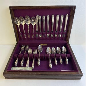 May include: A wooden cutlery chest with a purple velvet interior containing a set of silver-toned spoons, forks, and knives. The case features a label reading WM. ROGERS & SON. The utensils are arranged in designated slots within the box.