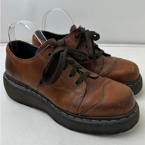 May include: A pair of brown leather oxford shoes featuring a rounded toe, dark brown laces, and a thick black rubber platform sole with yellow stitching detail along the welt. The shoes have a low-top design with visible stitching accents.