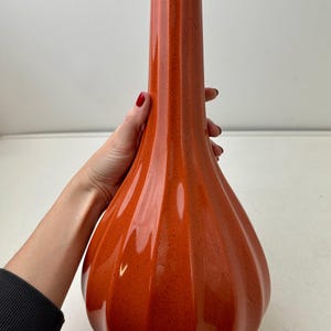 May include: A tall, orange ceramic vase with a speckled finish. The vase has a bulbous base and a fluted, cylindrical neck. The vase is approximately 12 inches tall.