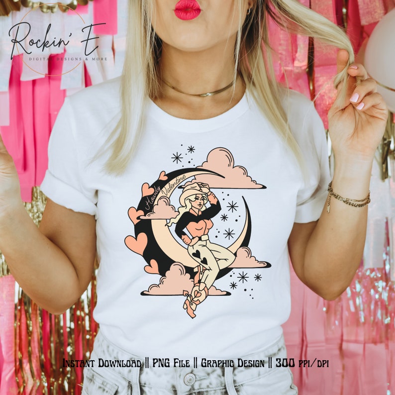Puede incluir: Una camiseta blanca con una ilustraci&oacute;n gr&aacute;fica de una persona con sombrero de vaquero sentada sobre una luna creciente. El dise&ntilde;o incluye nubes rosas, corazones negros, el texto Howdy Valentine y peque&ntilde;os detalles de estrellas.
