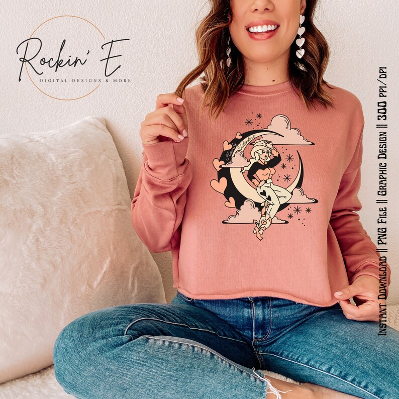 Puede incluir: Una sudadera de manga larga color rosa empolvado con dobladillo sin rematar que presenta un estampado gr&aacute;fico de una vaquera sentada sobre una luna creciente rodeada de corazones, nubes y estrellas. Incluye el texto "Howdy Valentine" y "Rockin' E".