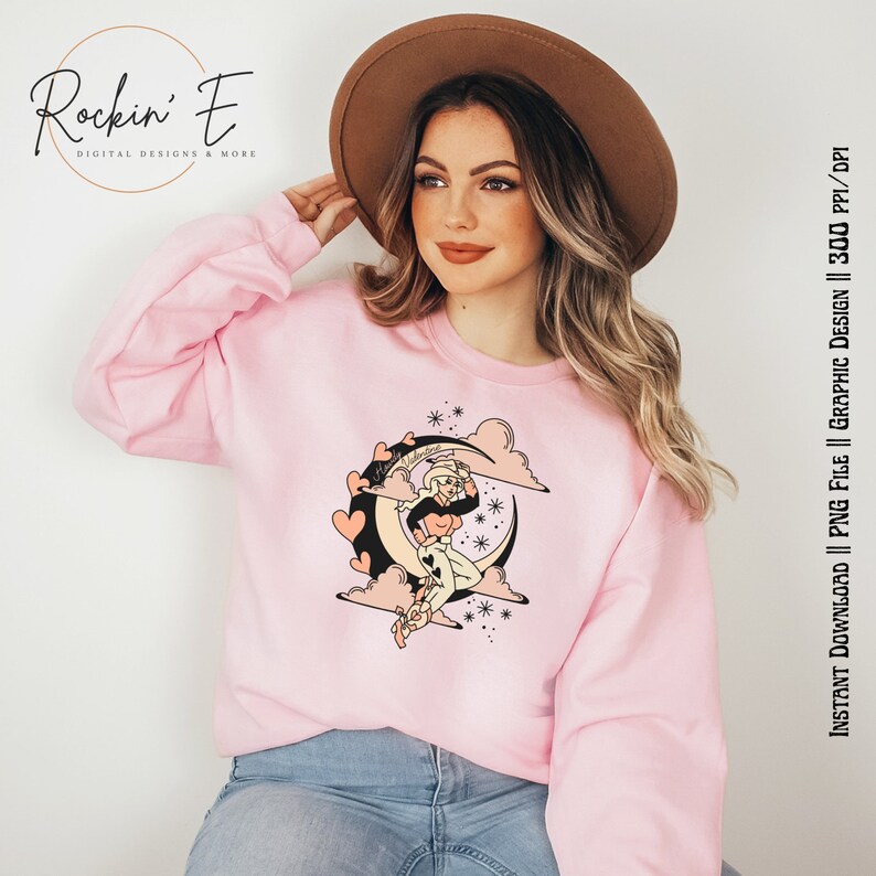 Puede incluir: Una sudadera de cuello redondo rosa claro con un estampado gr&aacute;fico de una vaquera sentada sobre una luna creciente rodeada de nubes, estrellas y corazones. El gr&aacute;fico incluye el texto "Happy Valentine" escrito en letra cursiva.