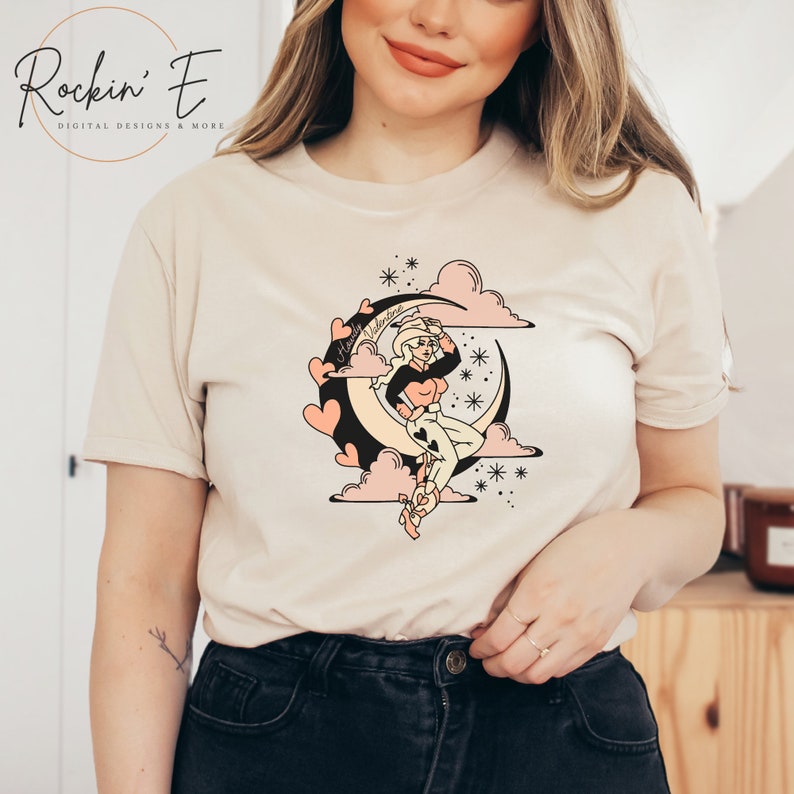 Puede incluir: Una camiseta beige de manga corta con un estampado gr&aacute;fico de una persona con sombrero de vaquero sentada sobre una luna creciente. El dise&ntilde;o incluye nubes rosas, corazones, estrellas y el texto Howdy Valentine en letra cursiva.