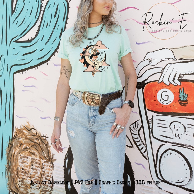 Puede incluir: Una camiseta de manga corta en color verde menta claro con un estampado gr&aacute;fico de una vaquera sentada sobre una luna creciente rodeada de nubes rosas y estrellas. La prenda se combina con vaqueros azul claro desgastados y una hebilla de cintur&oacute;n dorada.