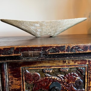 May include: A light beige, woven-textured decorative bowl with a unique, asymmetrical shape. The bowl is placed on a dark wooden surface. The background features a detailed, carved wooden cabinet with ornate designs.