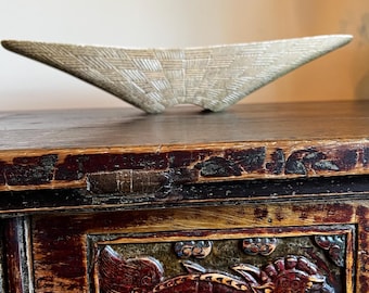 May include: A light beige, woven-textured decorative bowl with a unique, asymmetrical shape. The bowl is placed on a dark wooden surface. The background features a detailed, carved wooden cabinet with ornate designs.