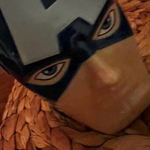 Puede incluir: Primer plano de una m&aacute;scara del Capit&aacute;n Am&eacute;rica, con un casco azul y blanco con la letra "A". La m&aacute;scara tiene dise&ntilde;os detallados de ojos y est&aacute; sobre un fondo marr&oacute;n texturizado.