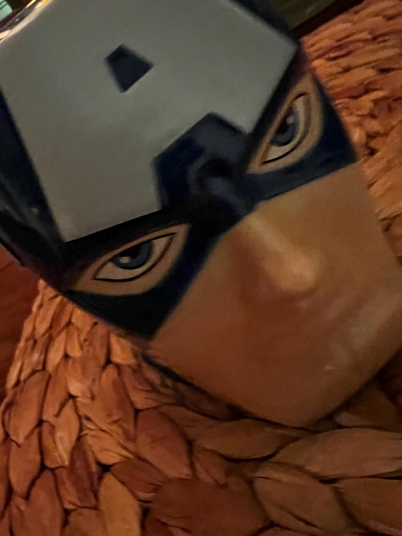 Puede incluir: Primer plano de una m&aacute;scara del Capit&aacute;n Am&eacute;rica, con un casco azul y blanco con la letra "A". La m&aacute;scara tiene dise&ntilde;os detallados de ojos y est&aacute; sobre un fondo marr&oacute;n texturizado.