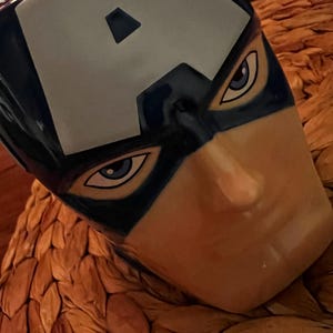 Puede incluir: Una taza de cer&aacute;mica con la cara del Capit&aacute;n Am&eacute;rica. La taza es principalmente azul con una "A" blanca en la frente. Los ojos est&aacute;n detallados con acentos azules y blancos, y la cara tiene un tono de piel bronceado. La taza est&aacute; sobre una superficie tejida y texturizada.