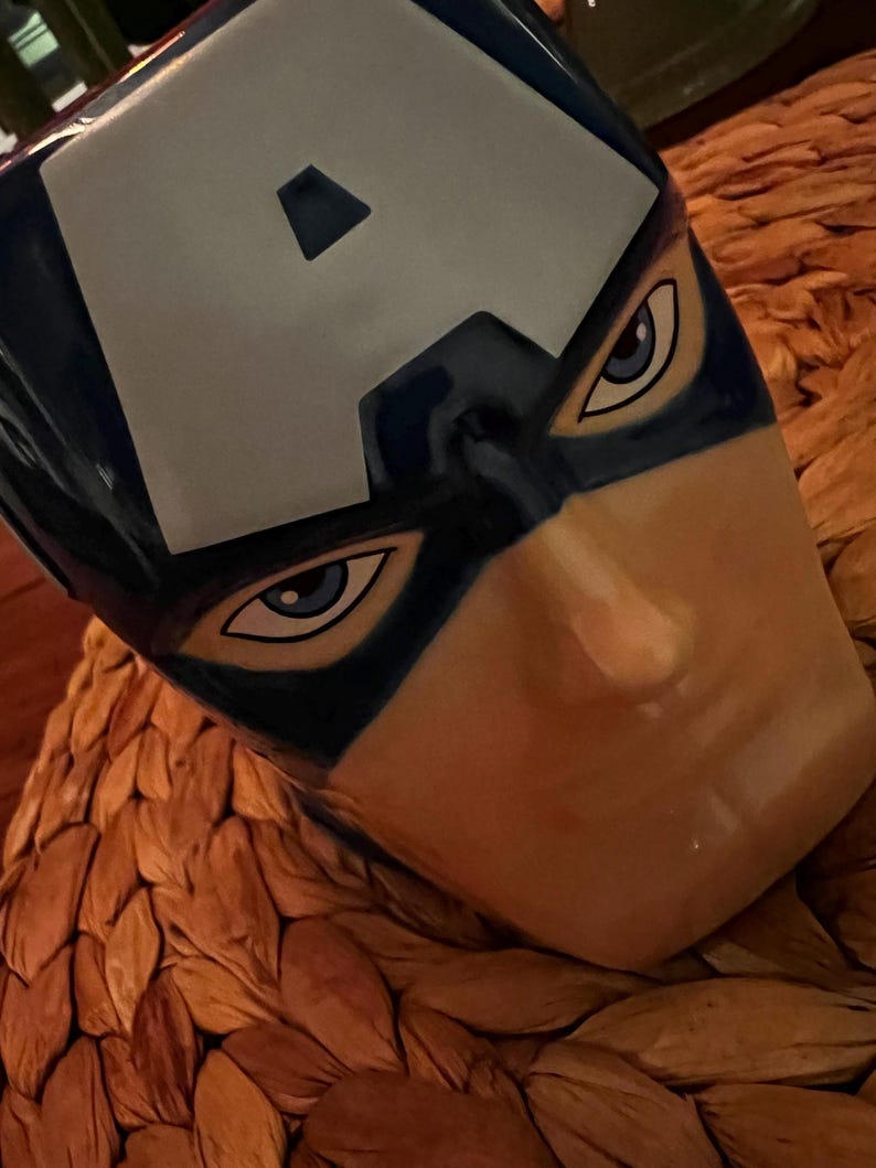 Puede incluir: Una taza de cer&aacute;mica con la cara del Capit&aacute;n Am&eacute;rica. La taza es principalmente azul con una "A" blanca en la frente. Los ojos est&aacute;n detallados con acentos azules y blancos, y la cara tiene un tono de piel bronceado. La taza est&aacute; sobre una superficie tejida y texturizada.