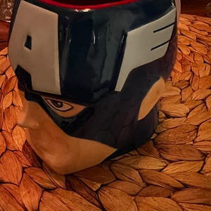 Puede incluir: Una taza de cer&aacute;mica con forma de la cabeza del Capit&aacute;n Am&eacute;rica. La taza presenta un casco azul con detalles blancos y la letra "A". El interior es rojo. La taza est&aacute; sobre un salvamanteles tejido.