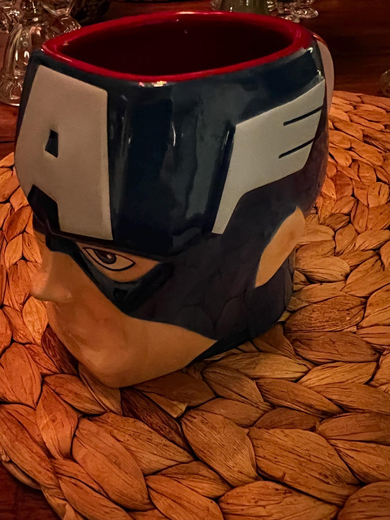 Puede incluir: Una taza de cer&aacute;mica con forma de la cabeza del Capit&aacute;n Am&eacute;rica. La taza presenta un casco azul con detalles blancos y la letra "A". El interior es rojo. La taza est&aacute; sobre un salvamanteles tejido.