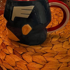 Puede incluir: Una taza de cer&aacute;mica azul marino con interior rojo y asa blanca y roja. La taza presenta un dise&ntilde;o blanco y gris en la parte delantera y un acento amarillo. La taza est&aacute; sobre un salvamanteles tejido de color beige.