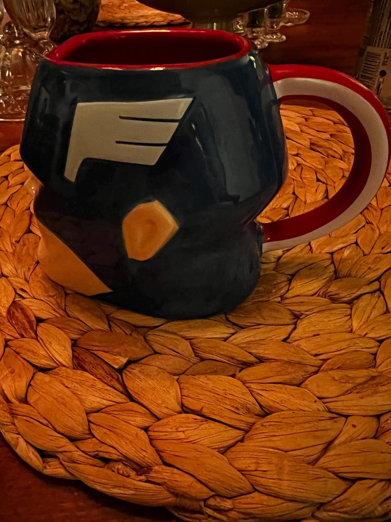 Puede incluir: Una taza de cer&aacute;mica azul marino con interior rojo y asa blanca y roja. La taza presenta un dise&ntilde;o blanco y gris en la parte delantera y un acento amarillo. La taza est&aacute; sobre un salvamanteles tejido de color beige.