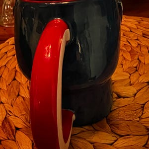 Puede incluir: Una taza de cer&aacute;mica con exterior azul marino e interior rojo. El asa es roja con una franja blanca. La taza se encuentra sobre una superficie tejida y texturizada. La taza es una pieza &uacute;nica para beber.